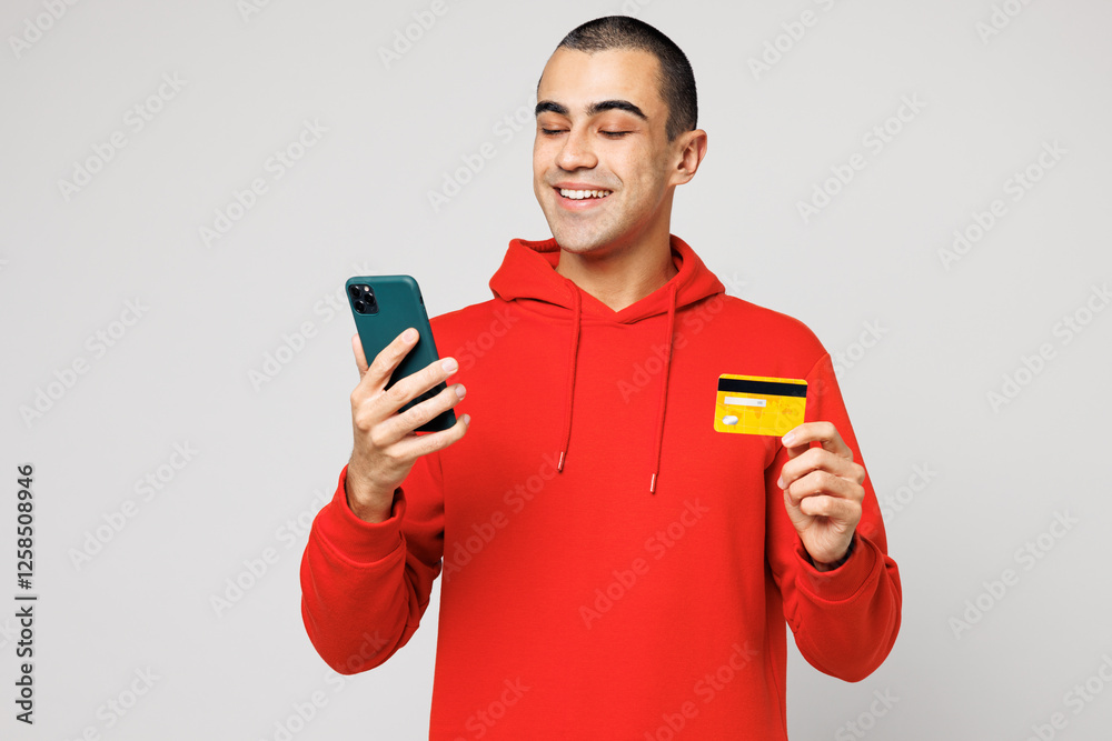 Young fun middle eastern man he wear red hoody casual clothes using mobile cell phone hold credit bank card doing online shopping order delivery booking tour isolated on plain solid white background.. Young fun middle eastern man he wear red hoody casual clothes using mobile cell phone hold credit bank card doing online shopping order delivery booking tour isolated on plain solid white background..