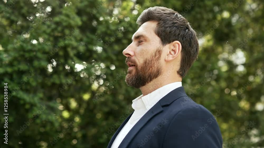Happy joyful businessman in a formal suit relaxing with closed eyes standing in urban city park. A smiling handsome bearded male breathes deeply, savoring fresh air in nature. Side view. Close up Happy joyful businessman in a formal suit relaxing with closed eyes standing in urban city park. A smiling handsome bearded male breathes deeply, savoring fresh air in nature. Side view. Close up