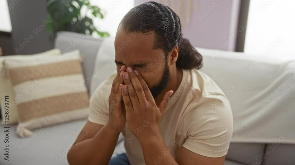 Young man with beard sitting in living room, appearing thoughtful and emotional in a cozy apartment setting with neutral decor. Young man with beard sitting in living room, appearing thoughtful and emotional in a cozy apartment setting with neutral decor.