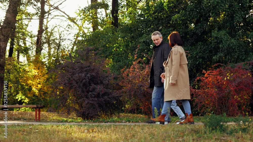 Family with son spends weekend together walking across fall botanical garden. Family of mom and dad leads little son by hands along path with bushes and bench. Family in warm clothes enjoy warm sunset Family with son spends weekend together walking across fall botanical garden. Family of mom and dad leads little son by hands along path with bushes and bench. Family in warm clothes enjoy warm sunset