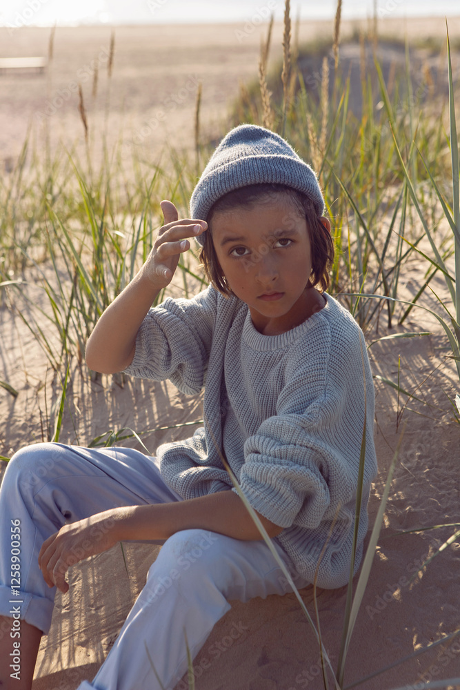 A fashionable seven-year-old boy sit along a sandy beach by the sea in autumn in a sweater and hat at sunset.