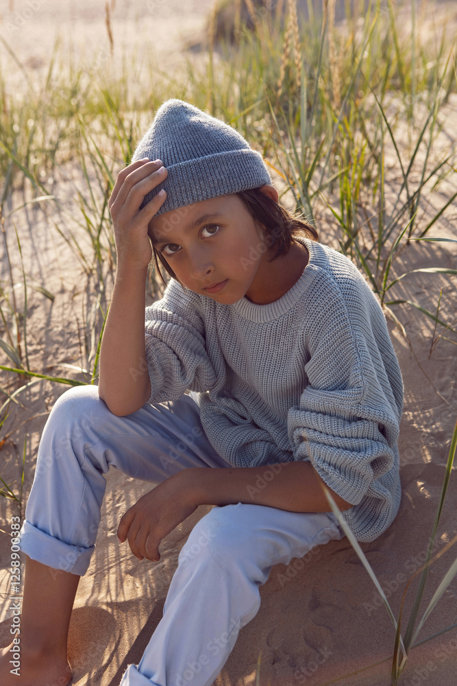 A fashionable seven-year-old boy sit along a sandy beach by the sea in autumn in a sweater and hat at sunset.