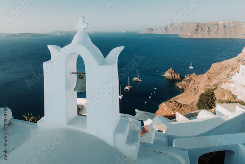 White bell tower with a bronze bell on an Oia street, volcanic cliffs. Thirasia in the background, and in the Aegean several ships sailing
