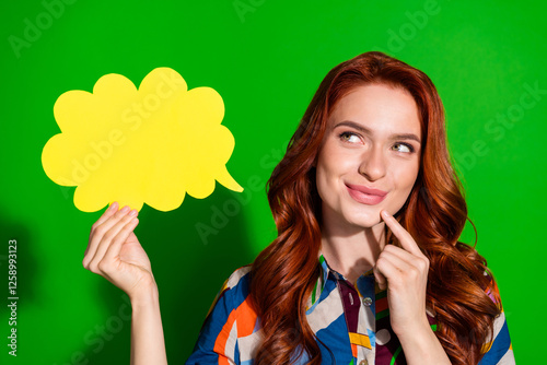 Young woman with red hair holding yellow thought bubble against green backdrop, wearing patterned shirt
