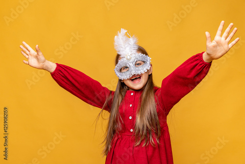 Overjoyed little girl wearing festive masquerade mask and red dress raises hands on yellow background. Child having fun at carnival party in Brazil or Venice. Carnival promotion and holidays concept photo