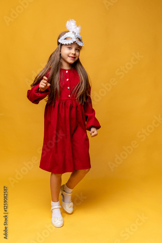 Overjoyed little girl wearing festive masquerade mask and red dress, dancing on yellow background. Child having fun at carnival party in Brazil or Venice. Carnival promotion and holidays concept photo