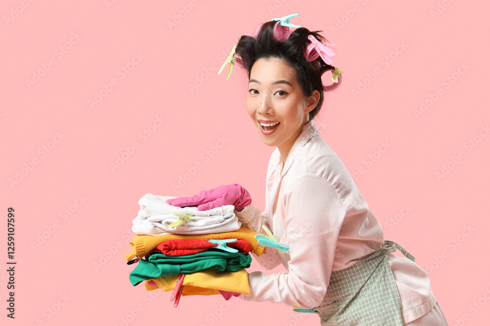 Happy young Asian woman in rubber gloves with stack of clothes and clothespins on pink background Happy young Asian woman in rubber gloves with stack of clothes and clothespins on pink background