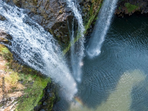 High-angle view of a powerful waterfall cascading down rocky cliffs into a pool below. Water splashes and a rainbow are visible. Whangarei Falls, Whangarei, Northland, New Zealand photo