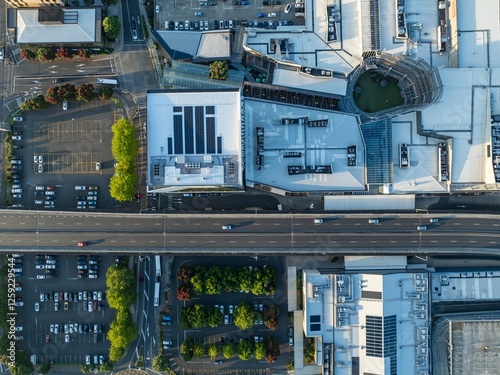 High-angle view of a bustling city shopping center. Cars park in lots, and a highway runs through the complex. Solar panels are visible on some buildings. SYLVIA PARK, AUCKLAND, NZ