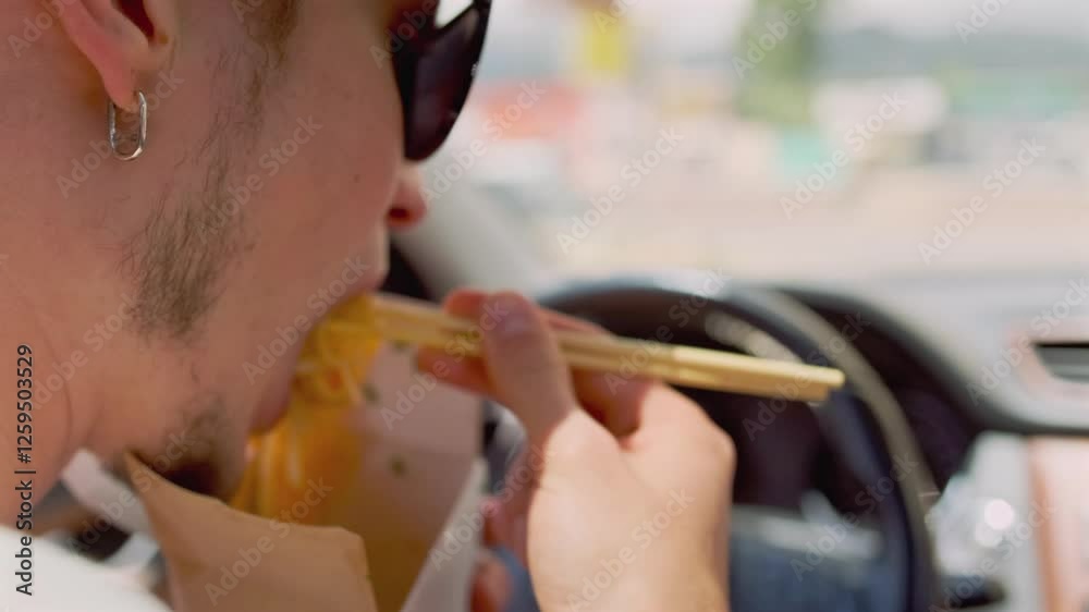 Side view. A man in sunglasses sits behind the wheel, eating udon wok noodles with meat and vegetables using chopsticks. Through the window, a bright summer day and a cityscape are visible.