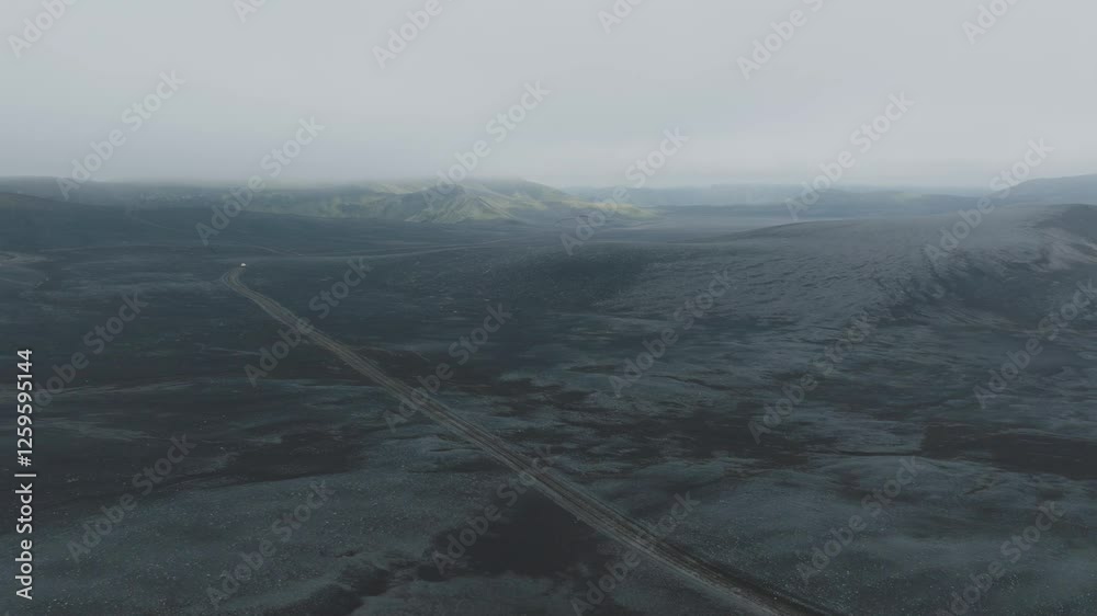 Aerial view of a lonely road cutting through vast Icelandic lava fields, surrounded by dark volcanic terrain. Hazy mountains in the distance add a mysterious atmosphere. Filmed at 50fps.