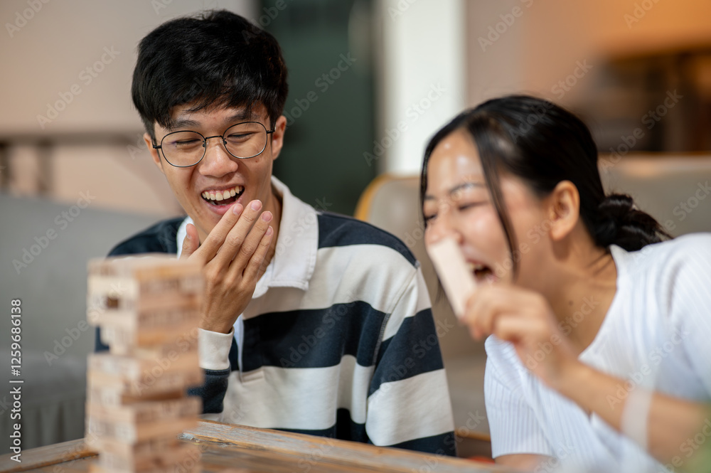 Two cheerful Asian friends, a male and a female, enjoy playing Jenga together at home. Two cheerful Asian friends, a male and a female, enjoy playing Jenga together at home.