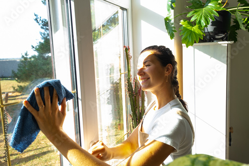 Woman in apron manually washes the window of house with rag cleaner and mop inside the interior with home plants on windowsill. Restoring order and cleanliness in the spring, cleaning servise photo