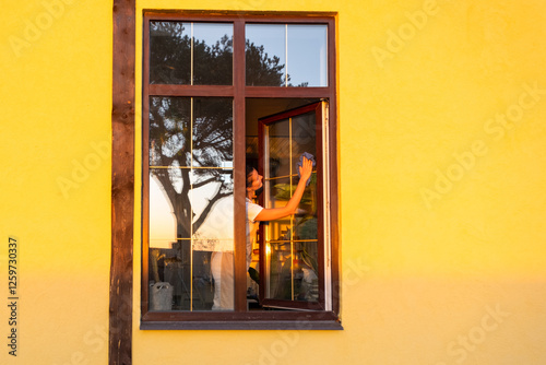 Woman in apron manually washes the window of the house with a rag with a spray cleaner and a mop outside. Safety at height, restoring order and cleanliness in the spring, cleaning service photo