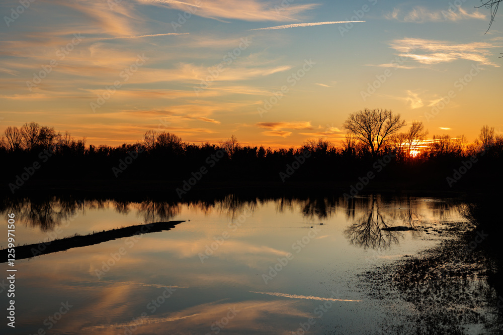 panoramica su un ambiente naturale fluviale al tramonto, con la luce solare che colora di arancione il cielo azzurro, leggermente nuvoloso, e si specchia sull'acqua ferma