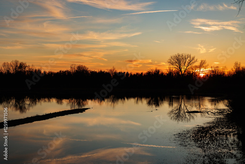 Obraz na płótnie panoramica su un ambiente naturale fluviale al tramonto, con la luce solare che colora di arancione il cielo azzurro, leggermente nuvoloso, e si specchia sull'acqua ferma w salonie