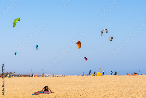 Kitesurfers and windsurfers preparing their kitesurfing equipment and flying kites over the beach in Tarifa, Spain, with the city and mountains in the background.
