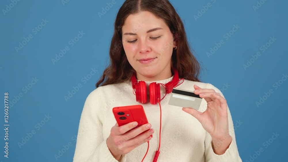 Focused young woman wearing red headphones around her neck, holding a credit card and smartphone, seemingly making an online payment or purchase. Blue background highlights the scene. Focused young woman wearing red headphones around her neck, holding a credit card and smartphone, seemingly making an online payment or purchase. Blue background highlights the scene.