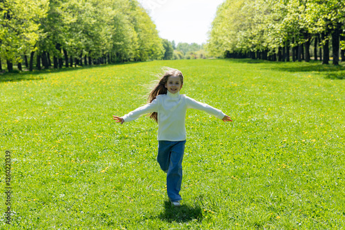 Little girl running across a green meadow in the park from afar towards the camera sunny day with outstretched arms. Freedom concept. Spring time and sunny day. Copy space photo