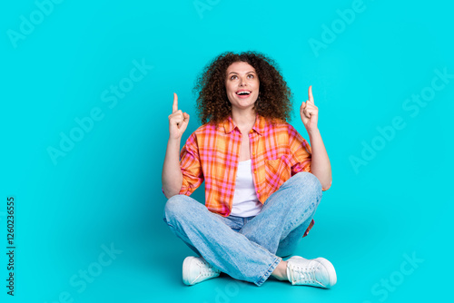 Young woman sitting cross-legged, pointing upwards with excitement on teal background, wearing a casual stylish outfit photo