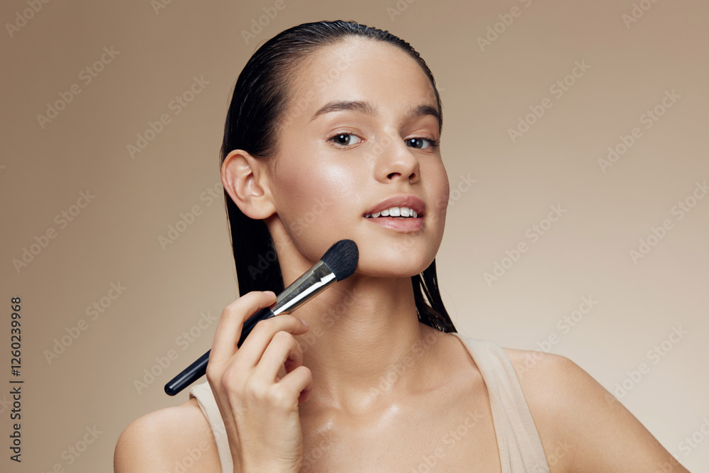 Young woman applying makeup with a foundation brush, showcasing a natural beauty look against a warm, neutral background Young woman applying makeup with a foundation brush, showcasing a natural beauty look against a warm, neutral background