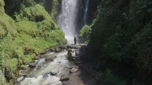 Verdant rainforest framing powerful waterfall cascading through Ecuadorian landscape, blonde female traveler overlooking pristine wilderness from aerial viewpoint