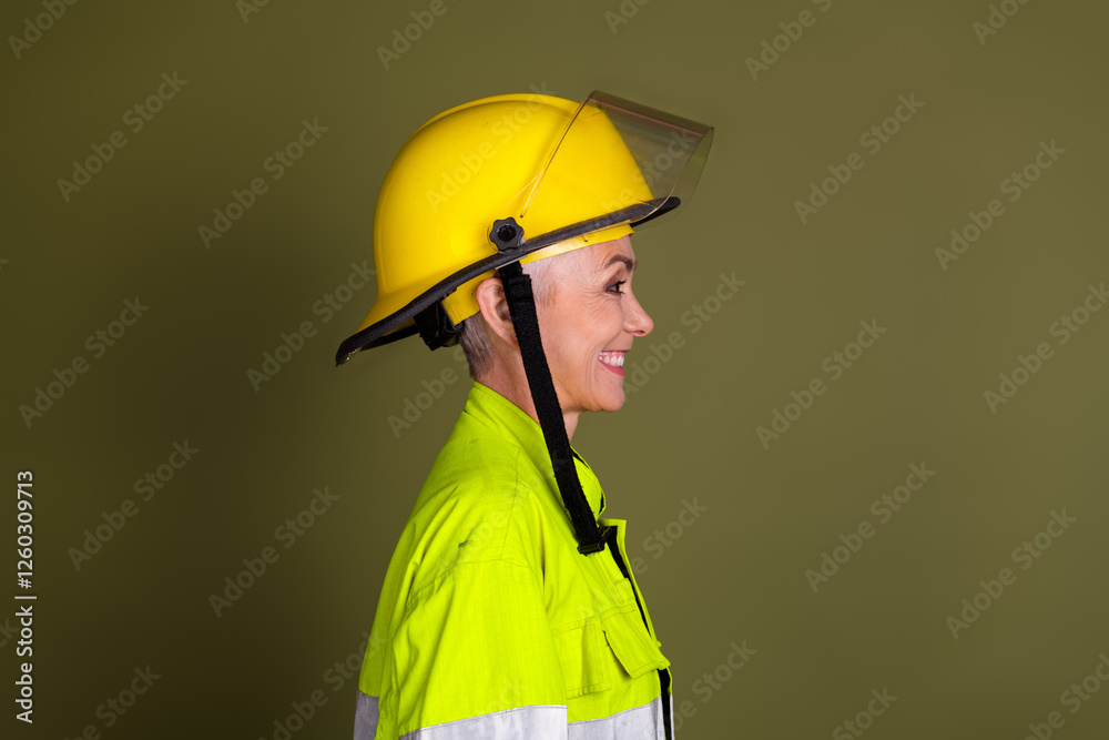 Profile view of a mature woman in high-visibility workwear and a helmet against a khaki background, emphasizing safety Profile view of a mature woman in high-visibility workwear and a helmet against a khaki background, emphasizing safety