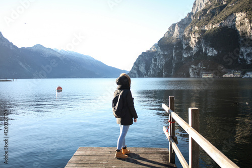 Photos A traveler in winter clothing stands on a wooden dock overlooking a serene mountain lake