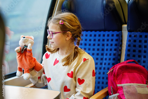 Obraz na płótnie Happy little girl with glasses and a colorful striped sweater sitting by a train window, holding a plush fox toy and looking outside with curiosity. Traveling child w salonie