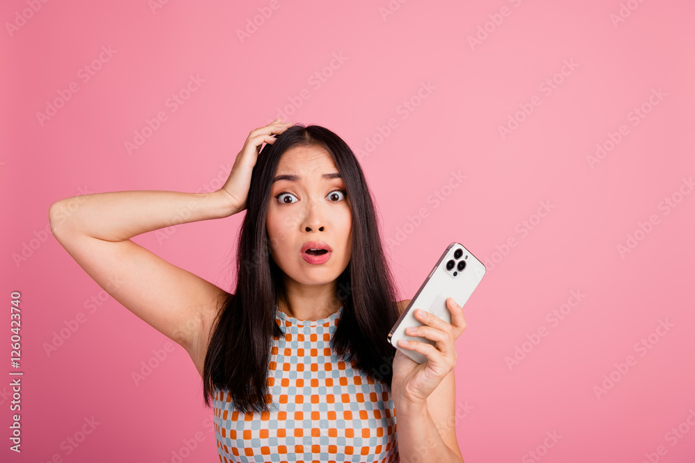 Naklejka premium Surprised young Asian woman with smartphone, wearing trendy top, in front of a pink background, experiencing shocking or unexpected news