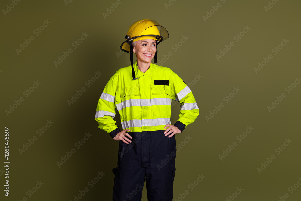 Mature female firefighter in reflective uniform and helmet stands confidently with hands on hips against khaki background Mature female firefighter in reflective uniform and helmet stands confidently with hands on hips against khaki background