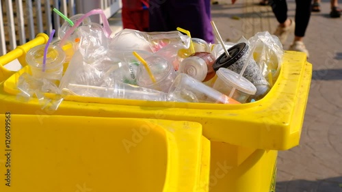 A yellow trash can is full of plastic cups and straws. The trash can is overflowing with plastic waste, and it is clear that people have been using disposable cups and straws