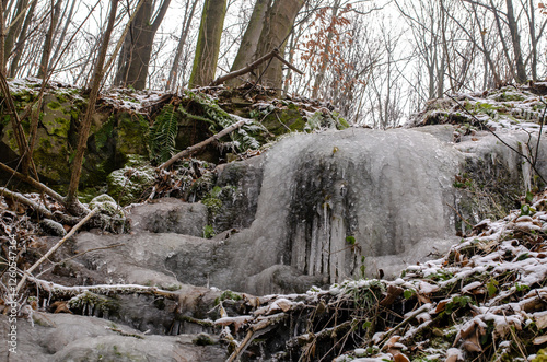 serene winter scene with a small frozen waterfall cascading over rocks covered in snow and moss, surrounded by a bare winter forest. The close-up view highlights the intricate details of the ice forma