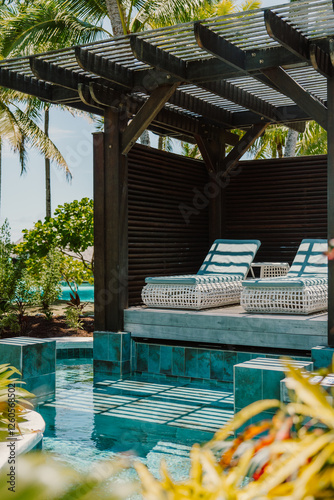 Fotografie Luxurious poolside cabana with elegant lounge chairs, shaded by a wooden pergola, surrounded by lush tropical greenery