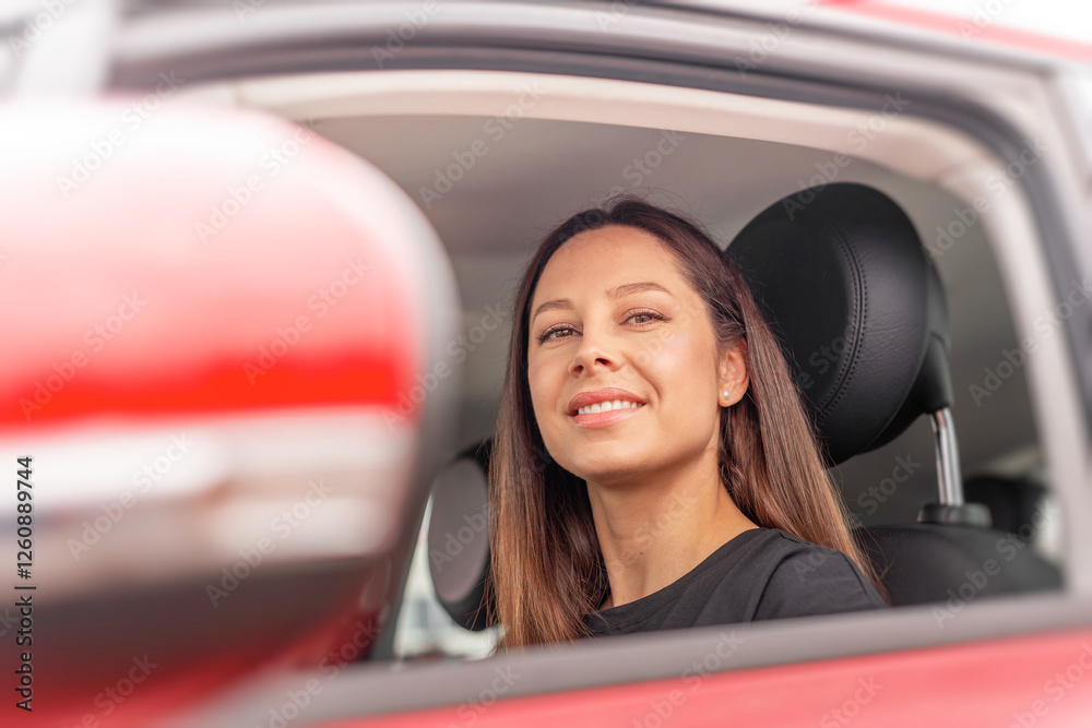 A young beautiful woman with long brown hair and a friendly smile sits in the drivers seat of a red car, looking outside the open window with a confident and relaxed expression.