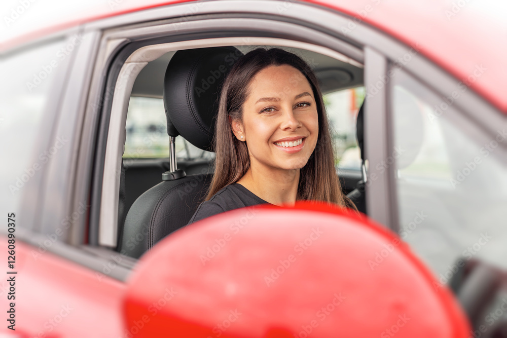 A young woman with long brown hair and a bright smile sits in the drivers seat of a red car, looking outside the open window with a cheerful and confident expression.