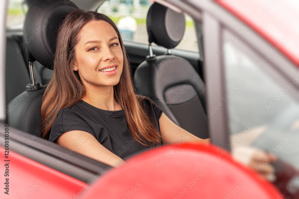 A young beautiful woman with long brown hair and a confident smile sits in the drivers seat of a red car, looking through the open window while holding the steering wheel.