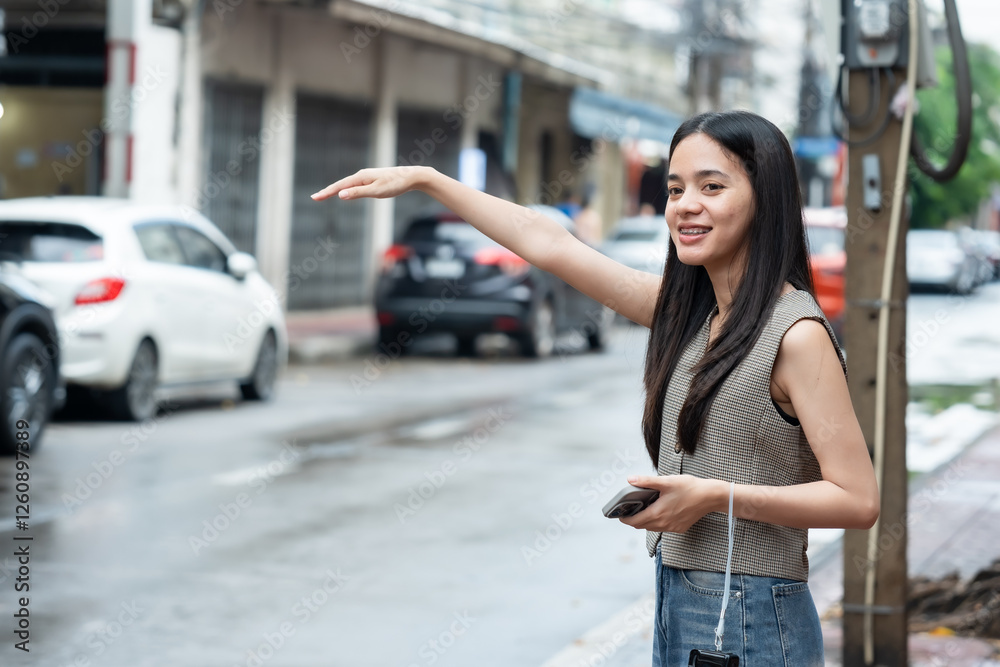 Fototapeta premium Beautiful Asian tourist walking in old city of Bangkok. Smiling young woman with long black hair waving hand to hail taxi. Holding smartphone and wearing stylish vest with casual jeans.