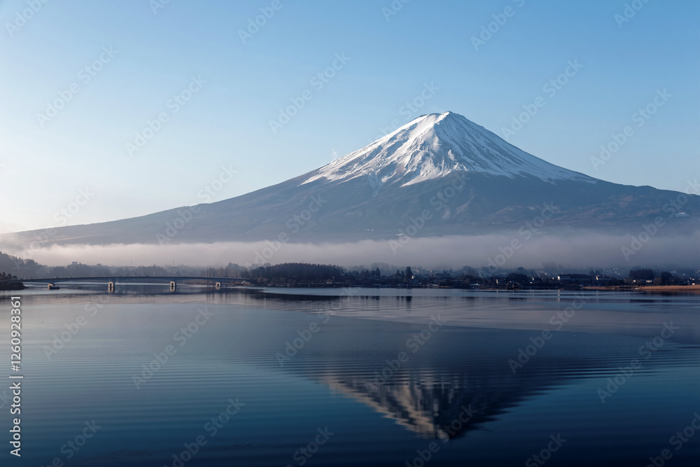 Scenery of snow capped Mount Fuji reflected in the peaceful water of Kawaguchi Lake with dense fog lingering at the foothill under blue clear sky on a sunny winter morning, in Yamanashi, Japan Scenery of snow capped Mount Fuji reflected in the peaceful water of Kawaguchi Lake with dense fog lingering at the foothill under blue clear sky on a sunny winter morning, in Yamanashi, Japan