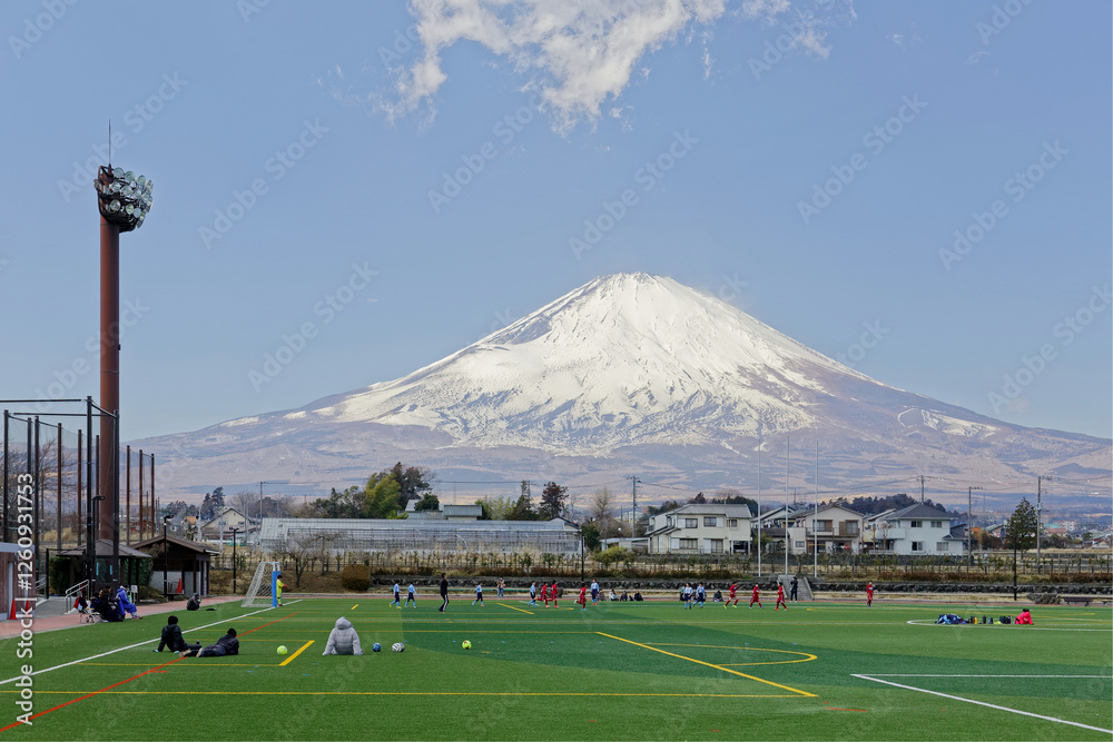 Fototapeta premium Young student-athletes exercising on a soccer field with the majestic snow capped Mount Fuji under blue sky in background on a sunny winter day, in the rural countryside of Gotenba, Shizuoka, Japan