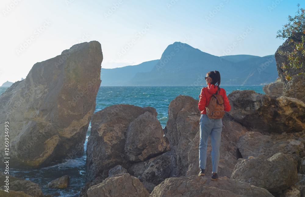Happy female tourist with a backpack on the top of a mountain, enjoying the sea view, standing on a rock and looking into the distance. The village of Novy Svet in Crimea. Russia