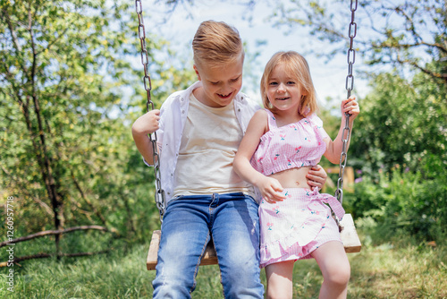 Blond boy and girl laugh and swing on swing. Joyful kids have fun outdoors in garden. Happy siblings are playing in park.