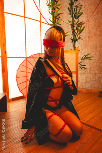 Sexy and beautiful young woman with blonde hair, black kimono and panties, and red shibari ties and ropes, red blindfold, holding a paper fan in a warm japanese room