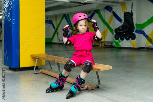 Little girl in pink suit sitting on a bench in skate park and puts on roller skates and protection before skating at rollerdrome. Sport and lifestyle concept. Copy space photo