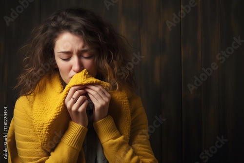 A woman with curly hair is wearing a bright yellow sweater, showing an emotional expression as she gently holds the fabric close to her face. The background features dark wood tones.