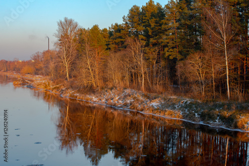 A serene river reflects a winter forest at sunset, with a light dusting of snow on the ground and a distant smokestack in the background.