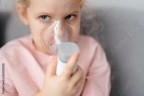 Portrait of blond child in inhalation mask with portable inhalation device with eyes close-up and steam. Childhood lung health.