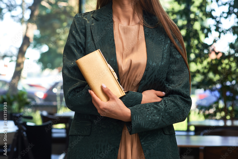 Elegant woman, gold clutch, green jacket, outdoor setting, refined fashion look, sophisticated style, secured wallet, day elegance, fashion focus concept