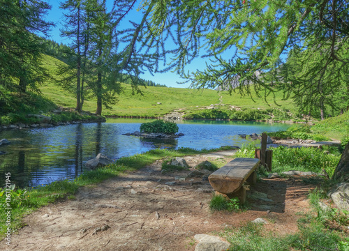Das Bild zeigt eine wunderschöne Landschaft einer bergigen Gegend mit einem kleinen See in der Mitte. Der See ist von grünen Bäumen und Sträuchern umgeben photo