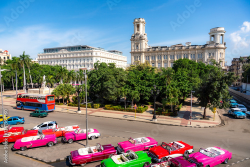 A row of vibrant classic convertible cars is parked near the Museum of the Revolution in Havana, Cuba, as seen from Parque Central, with lush greenery and colonial architecture in the background.   photo